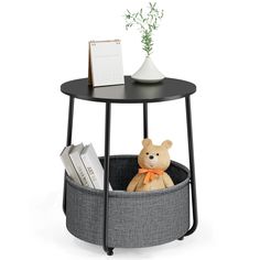 a teddy bear sitting in a grey basket under a table with books and a plant