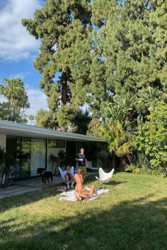 some people sitting on the grass in front of a house with trees and lawn chairs