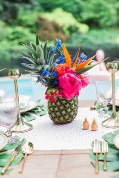 a pineapple centerpiece with colorful flowers and greenery sits on a table near a pool