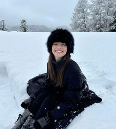 a woman sitting in the snow wearing a black coat and hat with fur on it