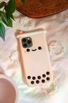 a white phone case with black dots and a smiling face on the back, sitting next to a plant