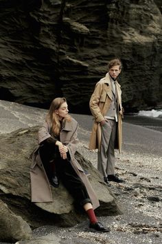 a man and woman are sitting on rocks near the ocean, one is wearing a trench coat