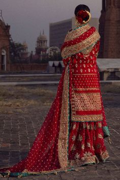 a woman in a red and gold bridal gown stands on a brick walkway with an old building in the background