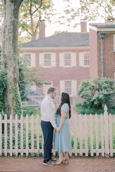 an engaged couple standing next to a white picket fence in front of a brick building