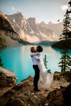 a bride and groom hugging on the edge of a cliff overlooking a mountain lake