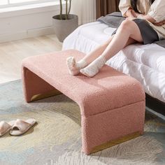 a woman sitting on top of a bed next to a pink foot stool with her legs crossed