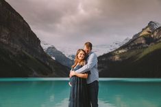 a man and woman standing next to each other in front of a mountain lake