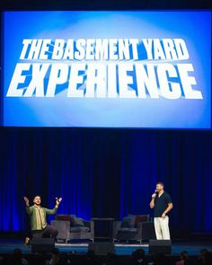 two men standing on stage in front of a large screen with the words basement yard experience