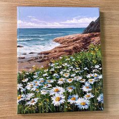 a painting of daisies by the ocean on a wooden surface with water in the background