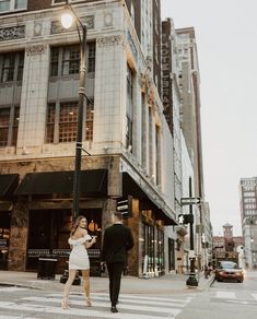 a man and woman crossing the street in front of a tall building at an intersection