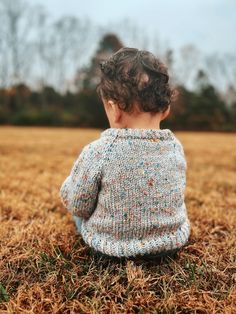 a small child sitting in the grass looking at something off to the side with trees in the background