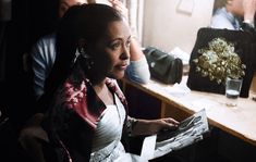 a woman sitting in front of a table holding a piece of paper and looking off into the distance