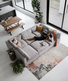 two people sitting on a large gray couch in the middle of a living room with lots of windows