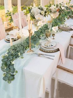 the table is set with white flowers and greenery on it, along with candles