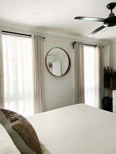 a white bed sitting under a ceiling fan in a bedroom next to a window with sheer curtains