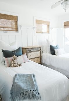 two twin beds with white linens and blue pillows in a small room that is decorated with wicker shutters