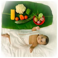 a baby laying on top of a bed next to a banana leaf and bowl of fruit