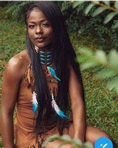 a woman with long black hair sitting on the ground in front of some trees and grass