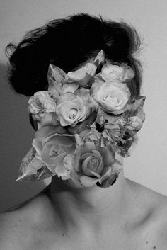 a black and white photo of a woman with flowers on her face, in front of a wall