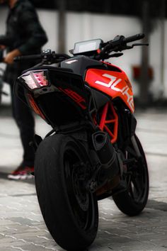 a red and black motorcycle parked next to a person on a sidewalk with another man in the background