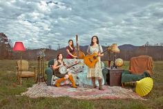 three women are sitting on a blanket in the middle of a field with guitars and umbrellas