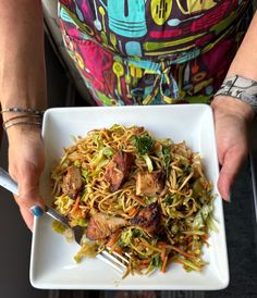 a woman holding a plate of food with meat and noodles on it, in front of her