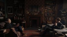 two men sitting on couches in a living room with bookshelves and fireplace