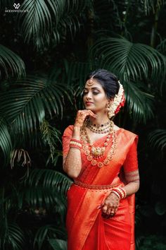 a woman in an orange sari and gold jewelry poses for the camera with palm trees behind her