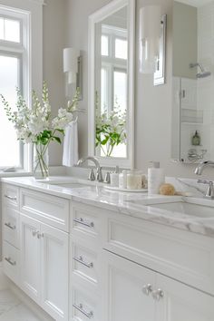 a white bathroom with double sinks and large mirrors on the wall next to two windows