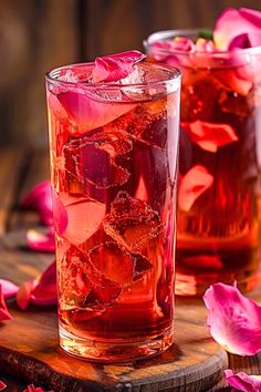 two glasses filled with ice and rose petals on top of a wooden board next to flowers