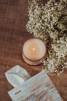 a white candle and some flowers on a wooden table next to tissue paper napkins