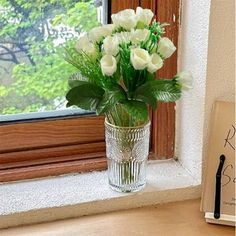 a glass vase with white roses in it on a window sill next to an open book