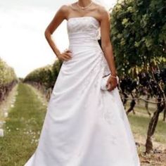 a woman in a white wedding dress standing next to a row of grape bushes and trees