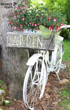 an old bicycle with flowers in the basket is parked next to a tree and has a sign that says garden on it