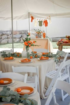 an outdoor tent set up with white chairs and orange plates on the table, along with greenery