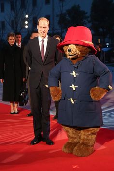 a man in a suit and red hat standing next to a large teddy bear on a red carpet