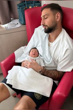 a man sitting in a red chair holding a baby while wearing a white shirt and black shorts