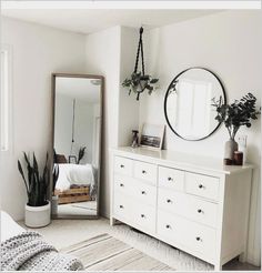 a bedroom with white furniture and plants on the dresser next to a large round mirror