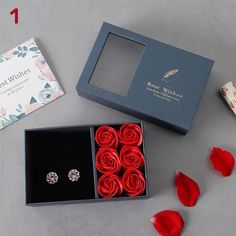 two red roses in a black box next to a pair of earrings and a card