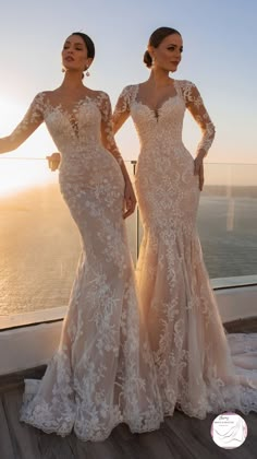 two women in wedding dresses standing next to each other on a balcony overlooking the ocean