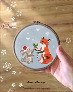 a hand holding a small cross stitched fox and bunny in front of snowflakes