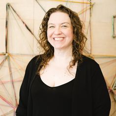 a woman with curly hair standing in front of a string art wall and smiling at the camera