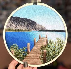 a person holding up a small embroidery art piece with a lake and dock in the background