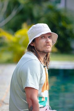 a man with long hair sitting next to a pool wearing a white hat and green shorts