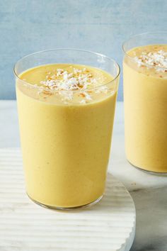 two glasses filled with yellow smoothie on top of a white plate and blue background