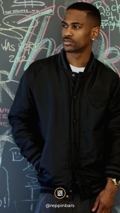 a man standing in front of a chalkboard with writing on it and wearing a black jacket