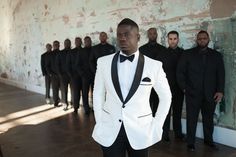 a man in a white tuxedo standing next to a group of black men