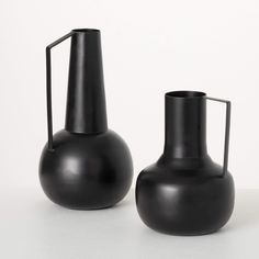 two black vases sitting next to each other on a white counter top, one is empty and the other has a handle