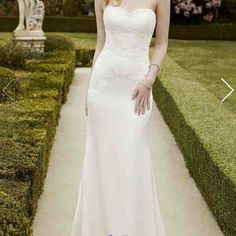 a woman in a white wedding dress standing on a path with hedges and bushes behind her