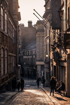 people are walking down the street in an old city at sunset or sunrise, with buildings lining both sides